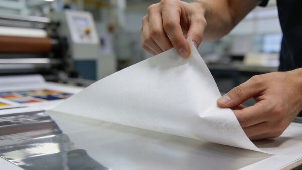 Silikonisiertes Trennpapier BRANOperl (Release Liner) wird kontrolliert und rückstandsfrei von einer industriellen Klebefolie gelöst. BRANOperl Silikonpapier für Druckereien, das in einem industriellen Produktionsumfeld von Hand abgezogen wird.