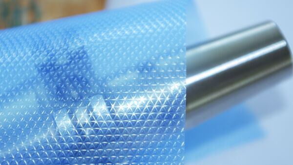 Nahaufnahme einer blauen BRANOpac VCI Prägefolie mit geprägter Oberflächenstruktur neben einer Metallrolle. Die VCI-Korrosionsschutzfolie schützt Metallteile und Metalloberflächen während Lagerung, Transport und Export vor Rost und Korrosion.