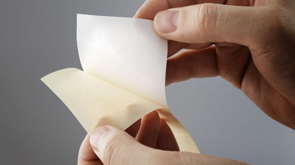 Hand löst ein weißes Trennpapier von einem silikonisierten Trägermaterial. Detailaufnahme eines einseitig beschichteten Trennpapiers für industrielle Anwendungen, Klebstoffe, Selbstklebetechnik und Verpackungslösungen.