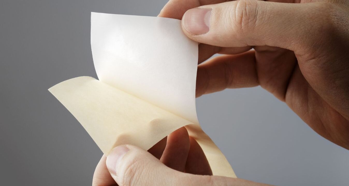 Hand löst ein weißes Trennpapier von einem silikonisierten Trägermaterial. Detailaufnahme eines einseitig beschichteten Trennpapiers für industrielle Anwendungen, Klebstoffe, Selbstklebetechnik und Verpackungslösungen.